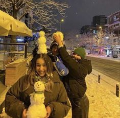 two people standing in the snow, one holding a snowman and smiling at the camera