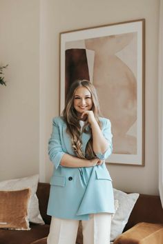 a woman standing in front of a couch wearing a blue blazer and white pants