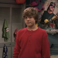 a young boy standing in front of a wall with pictures on it and wearing a red shirt