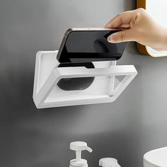 a person holding a cell phone in front of a wall mounted toilet paper dispenser