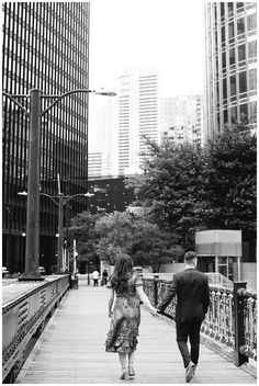 a man and woman walking down a bridge holding hands in the city with tall buildings behind them