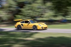 a yellow sports car driving down a road next to some grass and trees in the background