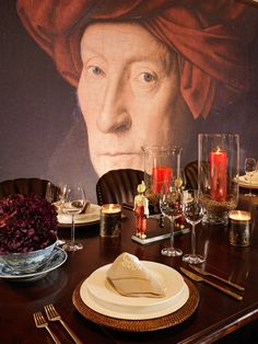 the table is set with plates, silverware, and candles in front of a painting of a man wearing a red turban