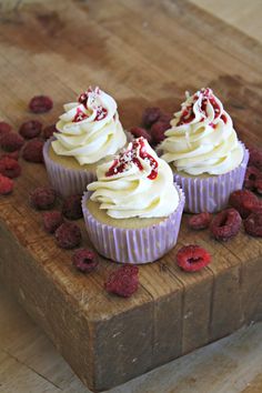 three cupcakes with white frosting and raspberries on a cutting board