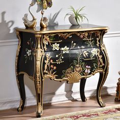 an ornate black and gold painted chest of drawers with flowers on the top, next to a potted plant