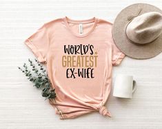 a pink shirt that says world's greatest ex - wife next to a hat