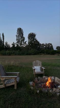 two wooden chairs sitting next to a fire pit in a field at dusk with trees and grass behind them