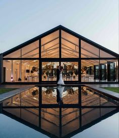 a bride and groom standing in front of a large glass building at dusk with their reflection on the water