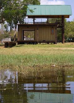 a small cabin sitting on top of a lush green field next to a lake in front of a forest