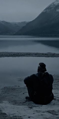 a person sitting on the ground looking out at water and mountains in the distance, with their back to the camera