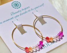 a pair of gold hoop earrings with multi colored beads on them sitting on top of a card