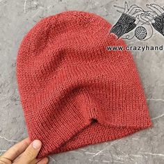 a hand is holding up a red knitted beanie on the concrete floor next to it