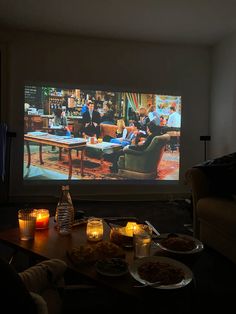 a couple of people sitting in front of a tv screen with food on the table