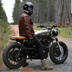 a man sitting on a motorcycle in the middle of a dirt road with trees behind him