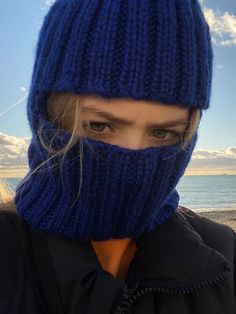 a woman wearing a blue knitted hat and scarf over her face with the ocean in the background