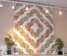 a large quilt hanging on the wall in a room with potted plants and lights