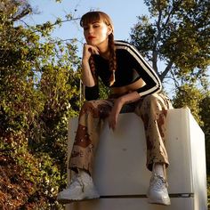 a woman sitting on top of a refrigerator in front of some trees with her hand under her chin