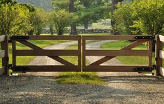 a wooden gate in the middle of a gravel road with trees and grass on either side