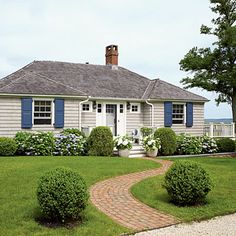 a white house with blue shutters and flowers in the front yard on a sunny day