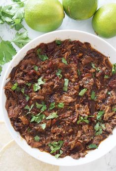 a white bowl filled with meat and garnish next to limes