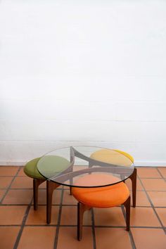 three tables sitting on top of a tiled floor next to a white wall and two orange stools