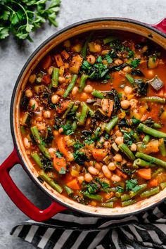 a red pot filled with beans and greens on top of a striped cloth next to a spoon