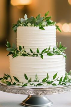 a white wedding cake with greenery on top is sitting on a silver platter