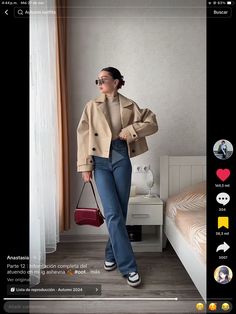 a woman standing in front of a bed wearing jeans and a trench coat, holding a handbag