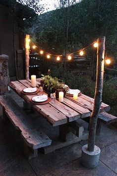 a wooden table with candles and plates on it in front of some lights hanging from the trees