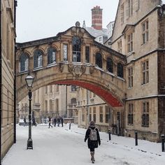 a woman walking down a snow covered street under an arched stone bridge in the middle of town