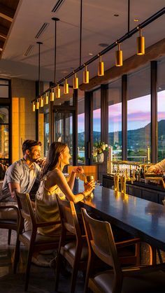 two people sitting at a bar with drinks in front of them and looking out the window