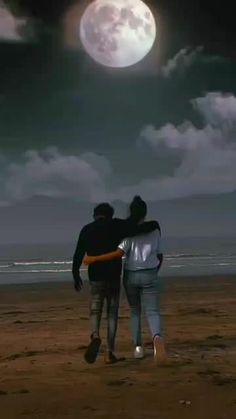 two people are walking on the beach at night with a full moon in the background