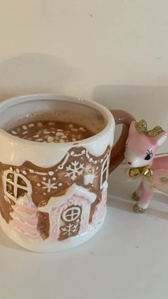 a white cup filled with hot chocolate next to a pink toy horse on top of a table