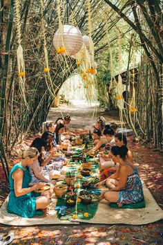 a group of women sitting around a table eating food in the woods with paper lanterns hanging overhead