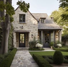 a stone house with hedges and trees in the front yard, along with white hydrangeas