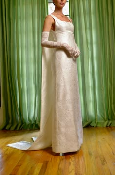 a woman in a white dress standing next to a window with green curtains behind her