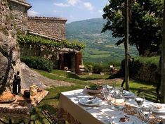 an outdoor table set with food and wine in front of a stone house overlooking the countryside