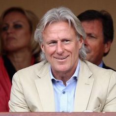 an older man in a white suit and blue shirt sitting at a table with other people behind him