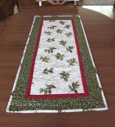 a christmas table runner with holly and berries on it sitting on top of a wooden table