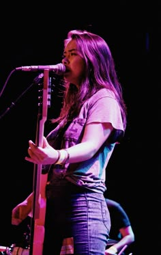 a woman standing in front of a microphone on top of a wooden stand and holding a guitar