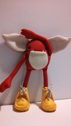 a red and white stuffed animal with yellow shoes on it's feet next to a wall