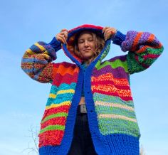 a woman wearing a multicolored knitted jacket and hoodie with her hands on her head