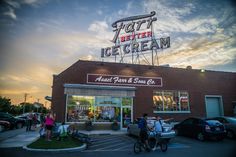 people are walking in front of the ice cream shop at sunset with cars parked outside