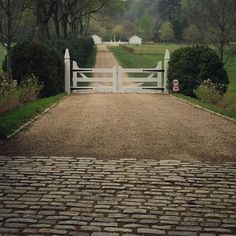 a white gate and brick walkway leading to a lush green field