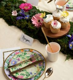 a table topped with plates and cups filled with cupcakes next to a moss covered wall