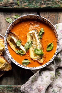 a bowl of tomato soup with bread and basil leaves on the table next to it