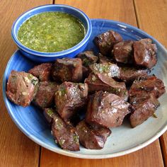 a blue plate topped with meat next to a bowl of green sauce on top of a wooden table