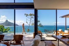 a woman walking past a living room filled with furniture and large windows overlooking the ocean