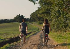 two people riding bikes down a dirt road next to a lush green field with trees