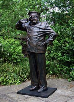 a statue of a man with a hat and jacket on standing in front of some bushes